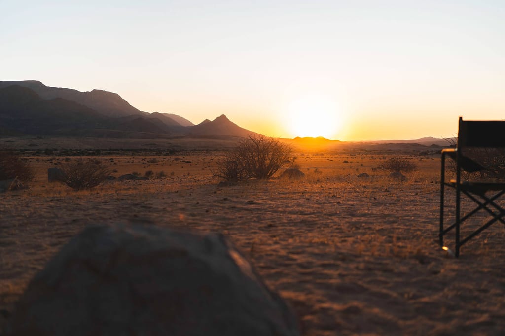 Weite, trockenes Wüstenland bei Sonnenuntergang mit Büschen, Bergen und einem Campingstuhl am Bildrand.