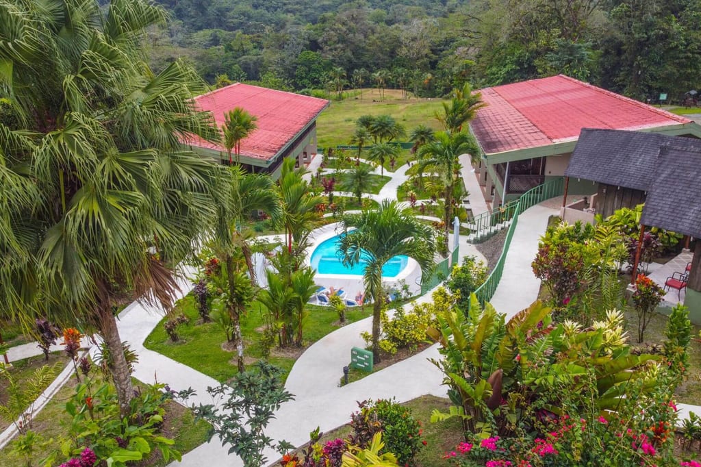 Vogelperspektive auf Resort mit Pool, Palmen, bunten Gärten und roten Dächern bei diffusem Tageslicht.