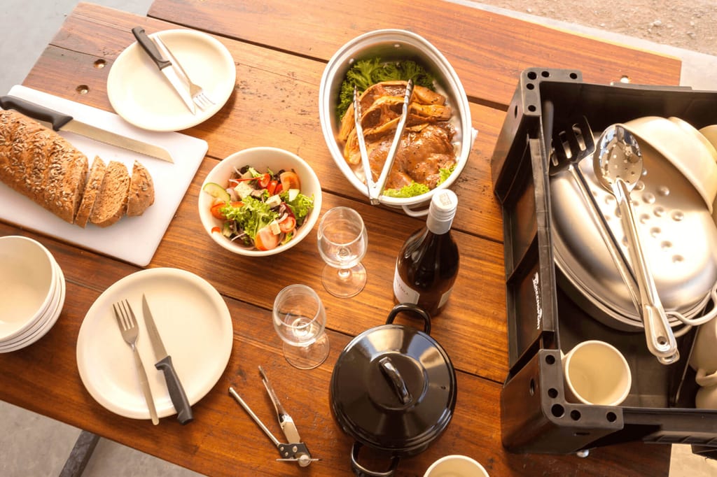 Gedeckter Holztisch mit Brot, Salat, Braten, Weinflasche und Geschirr im warmen Tageslicht