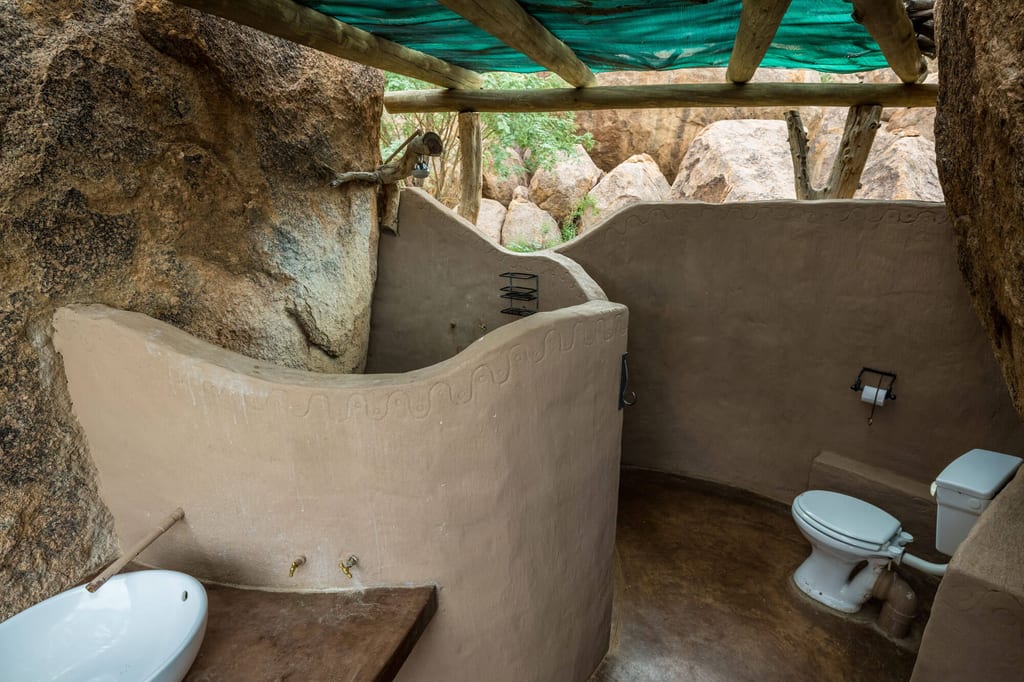Halb-offenes, rustikales Badezimmer in Felswand mit Toilette, Waschbecken und grüner Sonnenschutzplane