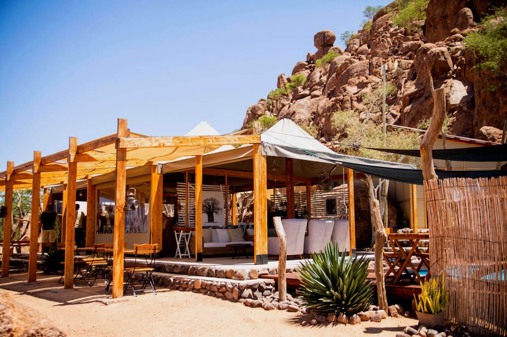 Freiluft-Lodge mit Holzpergola und offenen Zelten vor felsigem Hügel bei hellem Tageslicht.