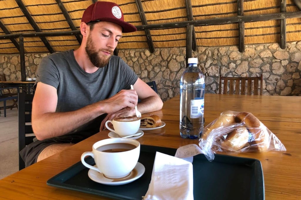 Mann rührt in einer Kaffeetasse an einem Holztisch mit Gebäck und Wasserflasche unter Strohdach.