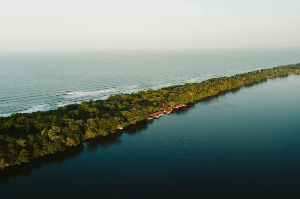 Luftbild einer schmalen, bewaldeten Landzunge zwischen ruhigem See und offenem Meer im warmen Abendlicht.