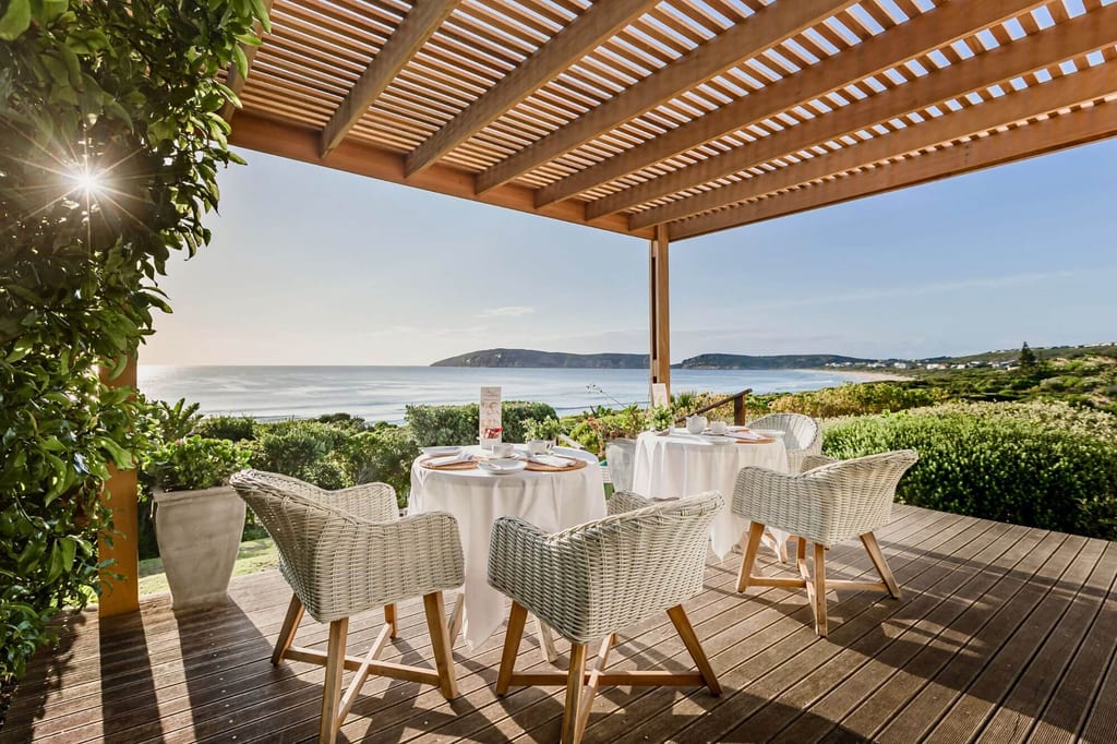 Überdachte Holzterrasse mit gedeckten Tischen und Korbsesseln vor Meer und Küstenlandschaft bei warmem Licht.