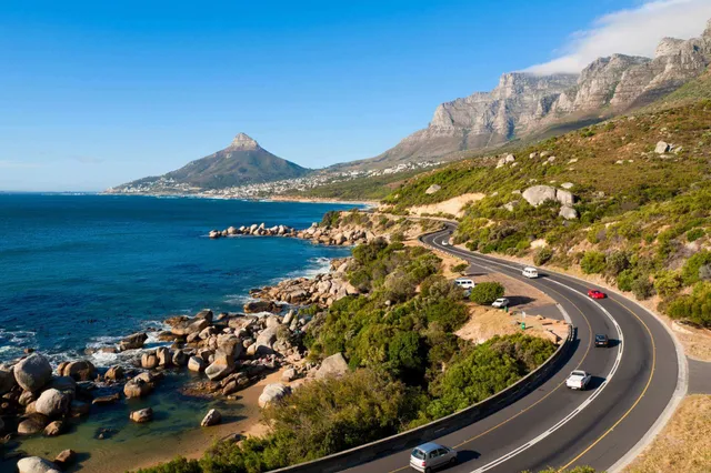 Erhöhter Blick auf eine geschwungene Küstenstraße in Südafrika mit Felsen, Meer und Autos bei Sonnenschein.