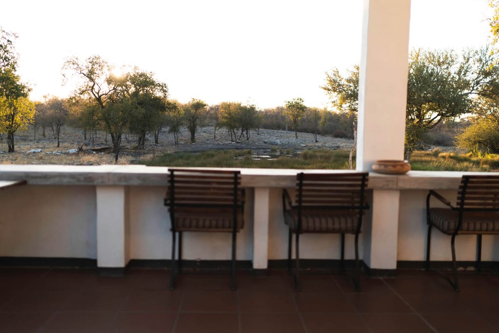 Terrasse mit drei Stühlen und Blick auf baumbestandene Landschaft und Wasserstelle im warmen Abendlicht.