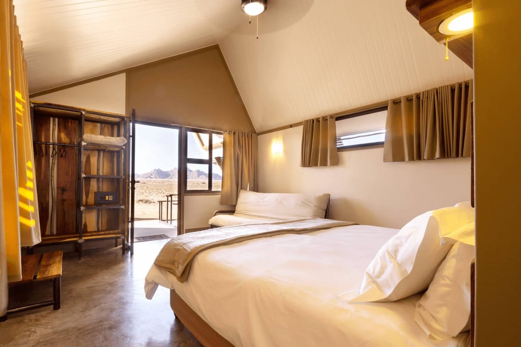 Gemütliches Lodge-Schlafzimmer mit offener Tür zur Wüstenlandschaft und Bergen am Horizont.