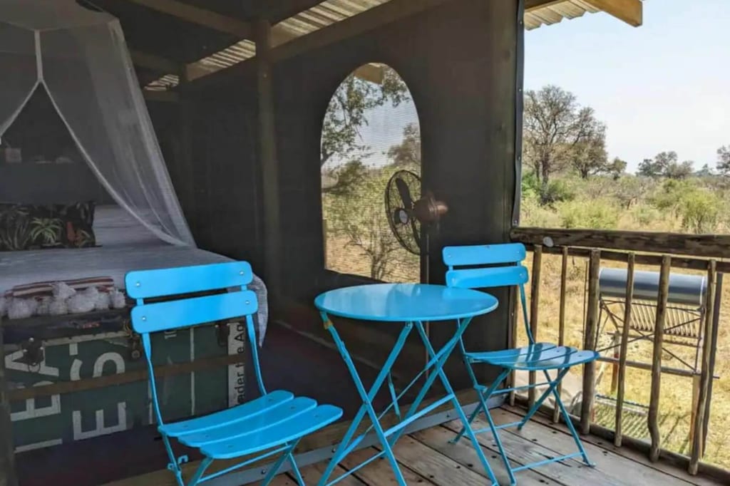 Veranda einer Safari-Zeltunterkunft mit türkisfarbenem Tisch und zwei Stühlen, Blick in die Buschlandschaft.