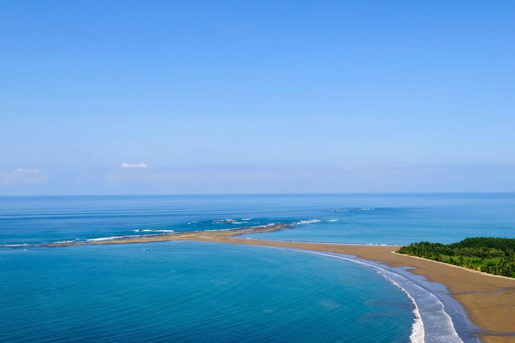 Luftaufnahme einer geschwungenen Sandbank und türkisblauem Meer mit bewaldeter Küste unter klarem Himmel.
