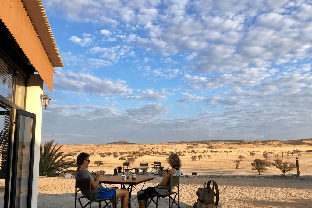 Zwei Menschen sitzen auf einer Terrasse und blicken in eine weite, sandige Halbwüste im warmen Abendlicht.