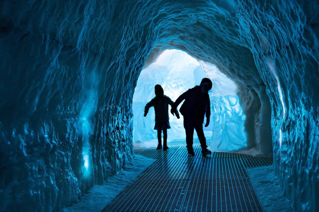 Blick durch einen blau beleuchteten Eistunnel mit zwei Personen als Silhouetten