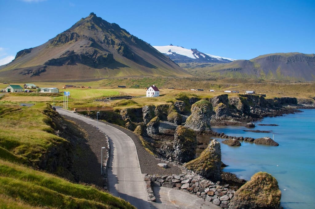 Küstenstraße entlang felsiger Klippen mit Felsstapeln, Dorf und Vulkanberg mit Gletscher im Hintergrund.