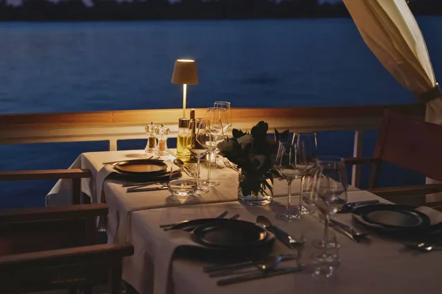 Festlich gedeckter Tisch auf einer Terrasse am Wasser bei Abenddämmerung mit warmem Lampenlicht.