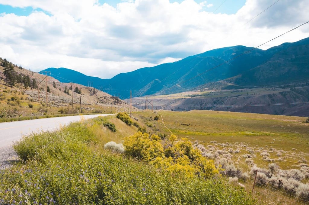 Weite Talansicht bei Cache Creek mit Landstraße, Büschen und Bergen unter wolkigem Himmel.