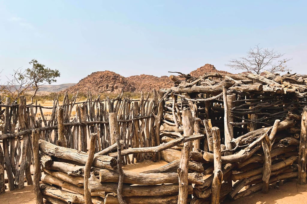 Rustikaler umzäunter Platz aus dicken Holzstämmen vor roten Felsen in trockener, sonnenbeschienener Landschaft.