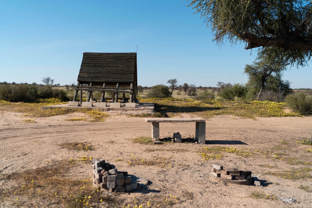 Sonnige Sandlandschaft mit erhöhtem Holzbau, Betonbank und kleinen Feuerstellen unter blauem Himmel.