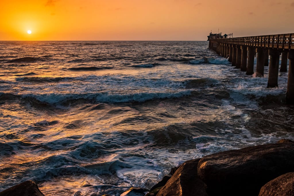 Sonnenuntergang über dem Meer mit Pier, brechenden Wellen und Felsen im Vordergrund.