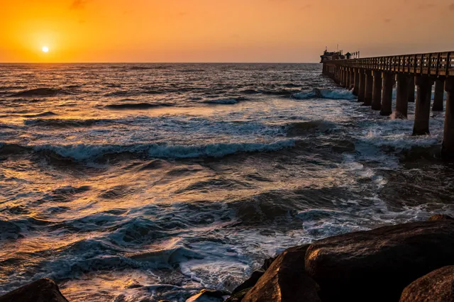 Sonnenuntergang über dem Meer mit Pier, brechenden Wellen und Felsen im Vordergrund.