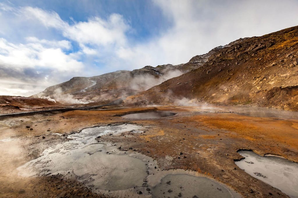 Geothermisches Feld mit dampfenden Schlamm- und Wasserpools vor kahlen Berghängen im diffusen Tageslicht.
