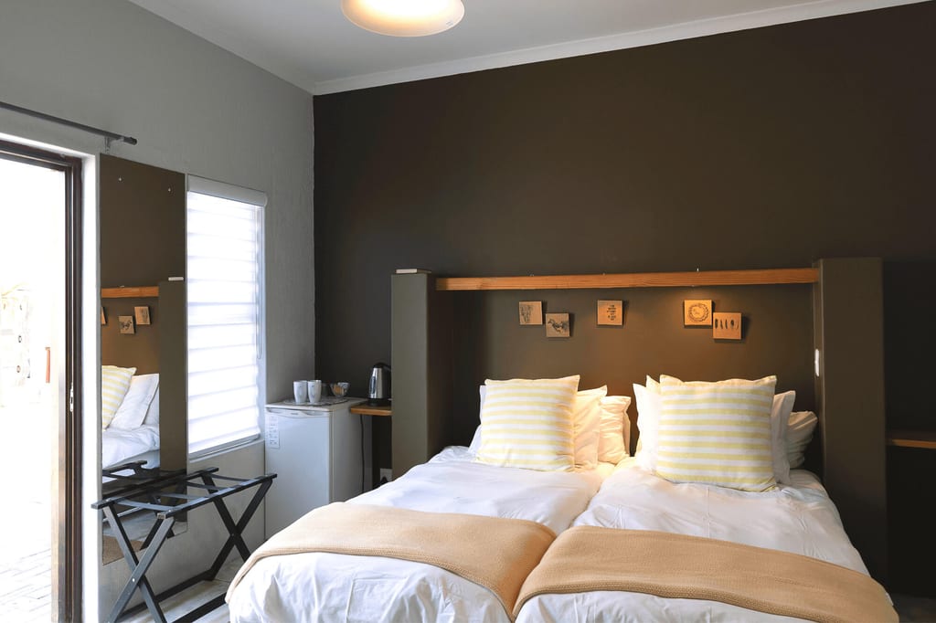 Zwei Einzelbetten mit hellen Kissen und Decken vor dunkler Wand, Fenster und Minibar links
