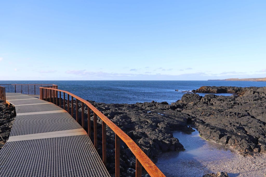 Metallsteg mit rostigem Geländer über schwarzer Lavaküste zum ruhigen blauen Meer unter klarem Himmel.