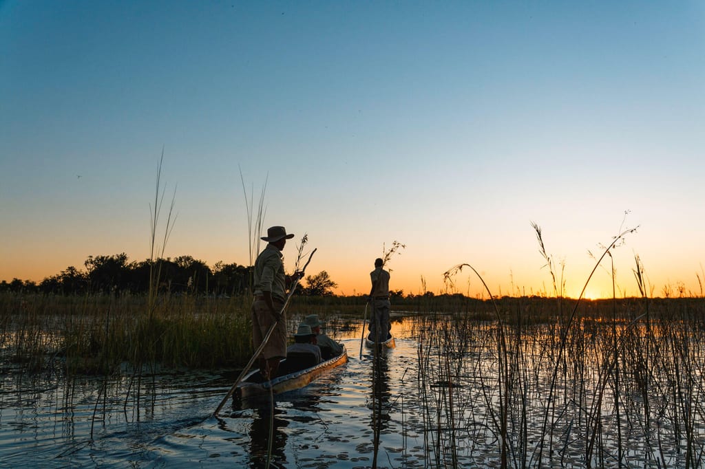 Zwei Personen polen mit flachen Booten durch Schilf bei Sonnenuntergang, ruhige Wasseroberfläche