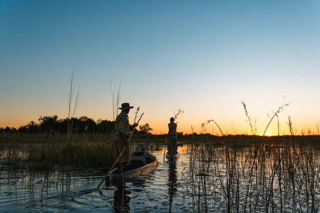Zwei Personen polen mit flachen Booten durch Schilf bei Sonnenuntergang, ruhige Wasseroberfläche