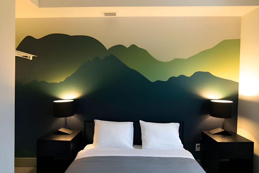 Schlafzimmer mit Doppelbett vor einer Wandmalerei aus stilisierten Bergen und zwei warm beleuchteten Nachttischlampen.