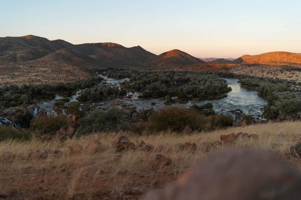 Panoramablick auf mäandrierenden Fluss mit Auenwald und Hügeln im warmen Abendlicht (Kaokoland, Namibia).
