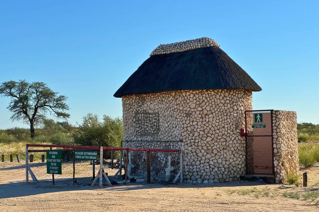 Runde Feldsteinhütte mit Reetdach auf sandigem Boden vor Savannenvegetation und klarem Himmel.