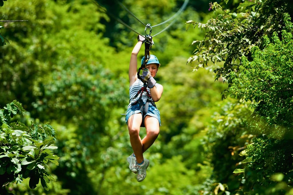 Person mit Helm und Gurt beim Zipline über grünen Wald an sonnigem Tag