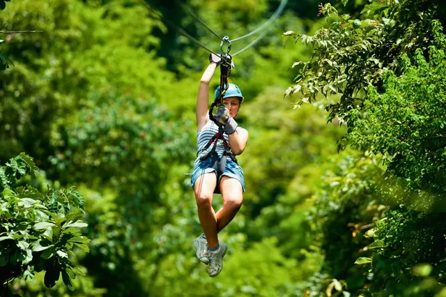 Person mit Helm und Gurt beim Zipline über grünen Wald an sonnigem Tag