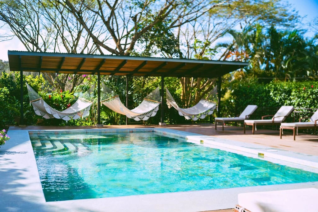 Sonniger Gartenpool mit Hängematten unter einer Pergola und Liegestühlen