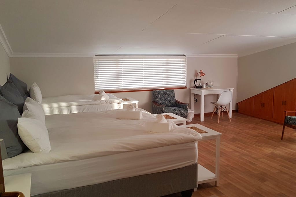 Helles Schlafzimmer mit zwei Betten, Schreibtisch und Sessel, weiches Tageslicht durch Jalousien