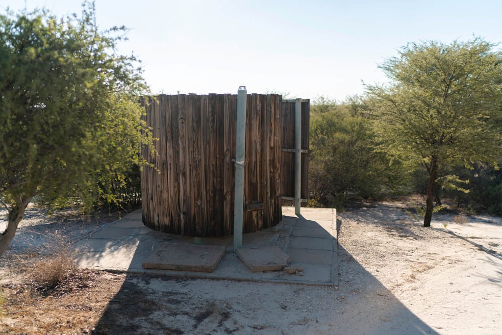 Runder hölzerner Sichtschutz auf Betonplatte in trockener, sandiger Buschlandschaft bei hellem Tageslicht.