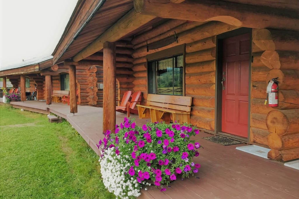 Veranda eines Blockhauses mit Holzbänken und einem Blumentopf mit lila und weißen Blüten.