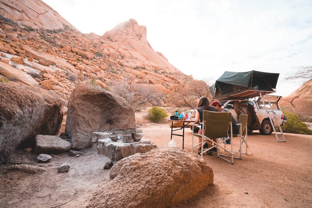 Zwei Personen beim Camping mit Dachzelt vor roten Felsformationen in einer Wüstenlandschaft.