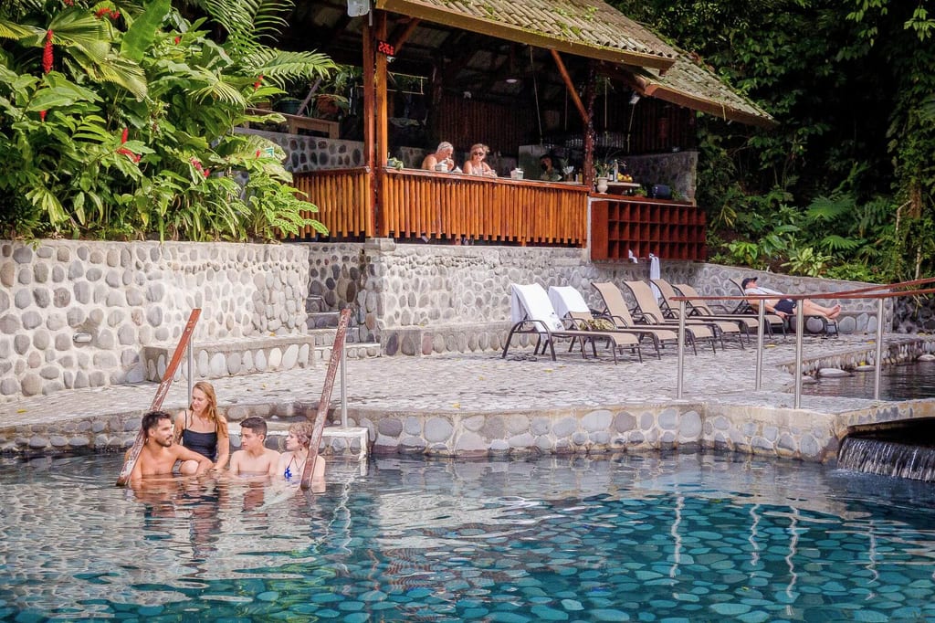 Vier Personen im Pool vor einer offenen Holzbar und Liegen, umgeben von tropischer Vegetation.
