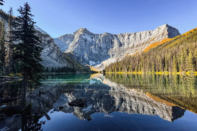 Bergsee mit spiegelglatten Wasser und reflektierenden Felswänden, Nadelwald am Ufer und blauem Himmel.