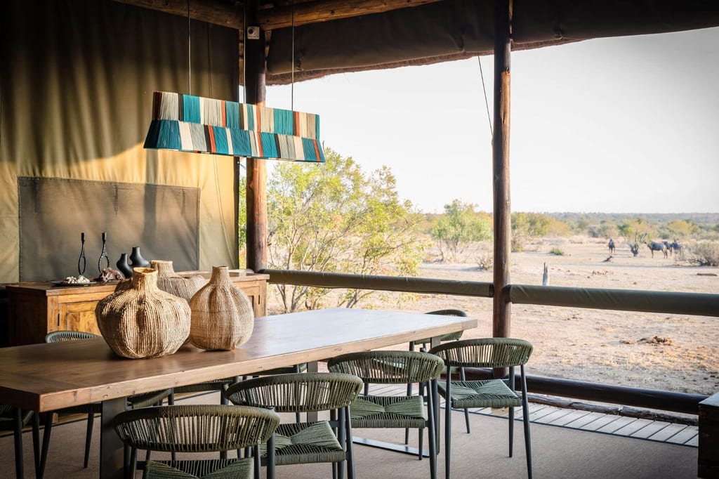 Offener Essbereich einer Safari-Lodge mit Holztisch und Blick auf trockene Savanne und Huftiere in der Ferne.