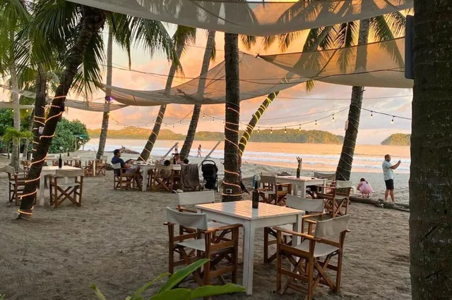 Strandrestaurant unter Palmen mit Tischen, Lichterketten und Abendsonne am Meer