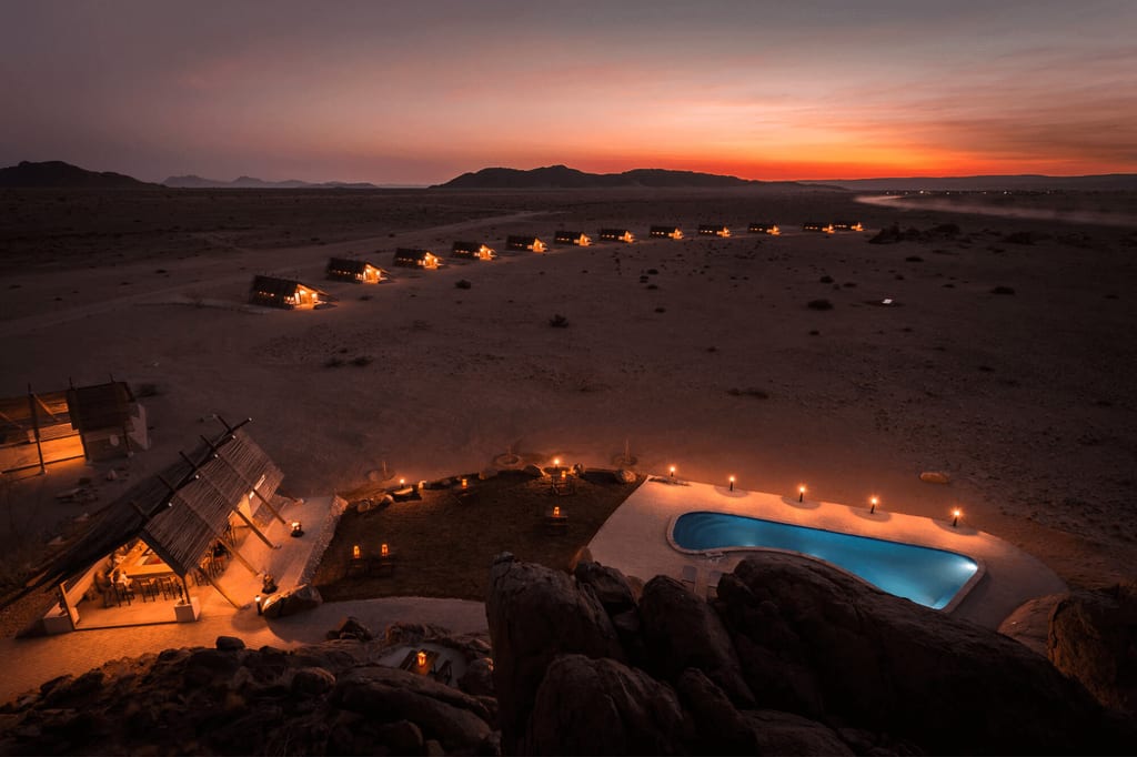 Abendliche Aufnahme eines beleuchteten Wüstenlagers mit Pool und Zeltreihe bei Sonnenuntergang.