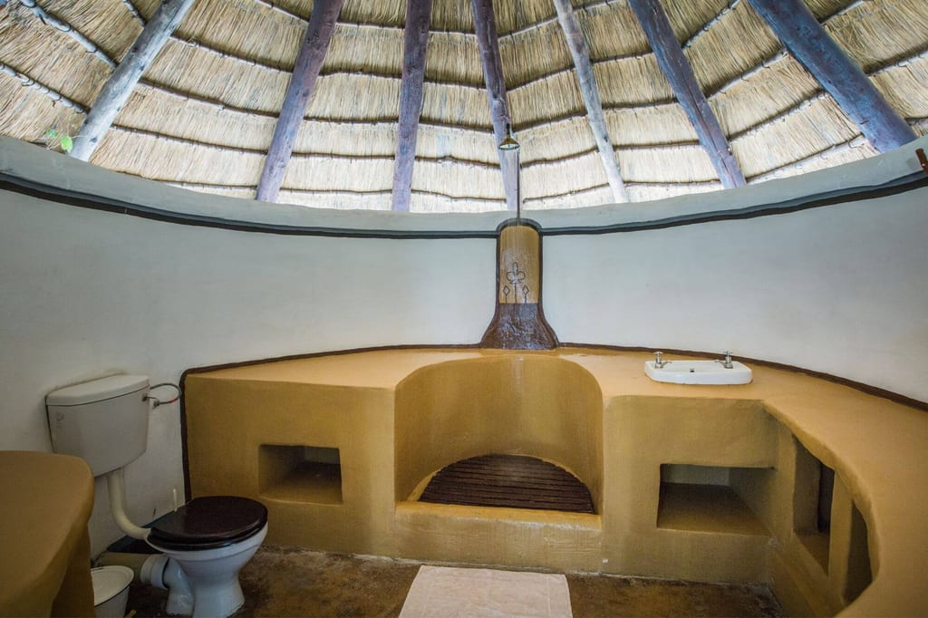 Rundes Badezimmer mit Strohdach, ockerfarbenen Einbauten und Toilette, frontal aufgenommen.