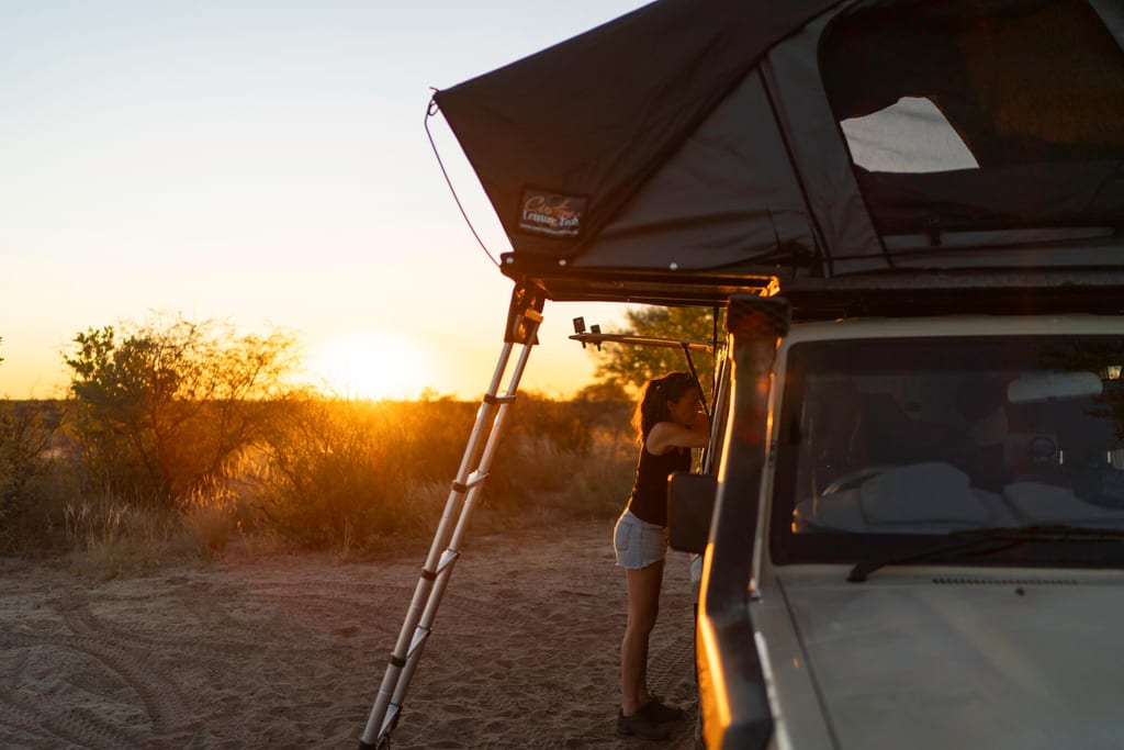 Frau neben Geländewagen mit Dachzelt bei Sonnenuntergang in trockener Buschlandschaft