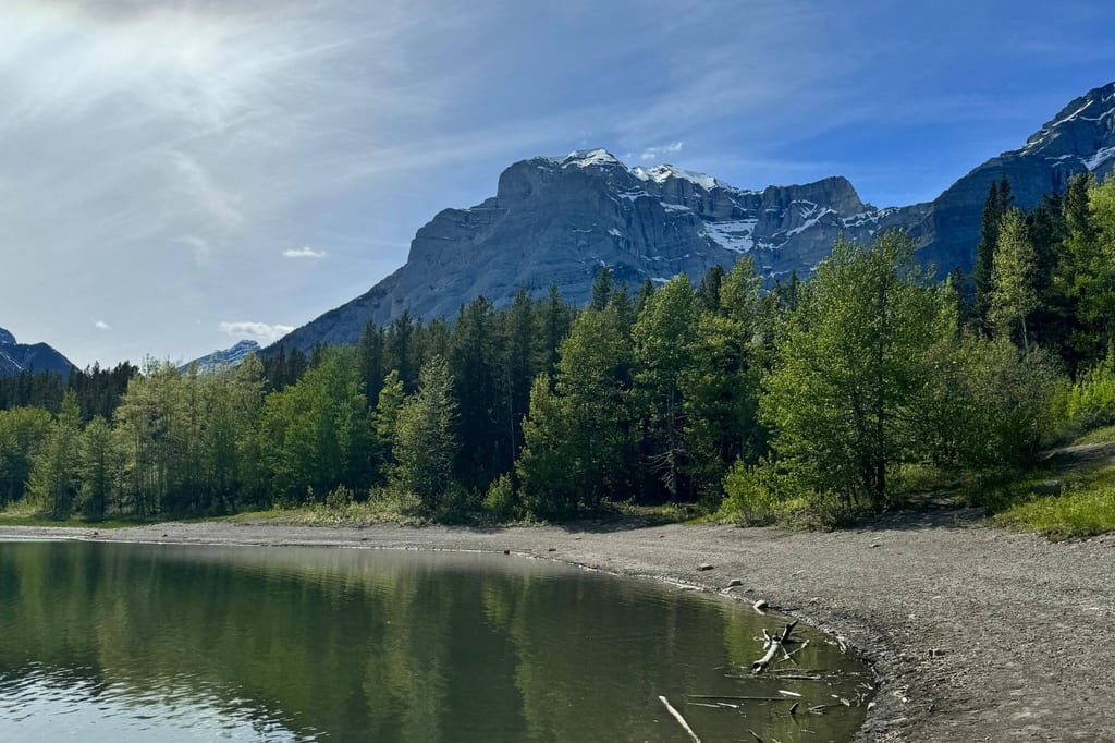 Bergsee mit Ufer und spiegelnden Bäumen vor schneebedeckten Felsen im klaren Nachmittagslicht