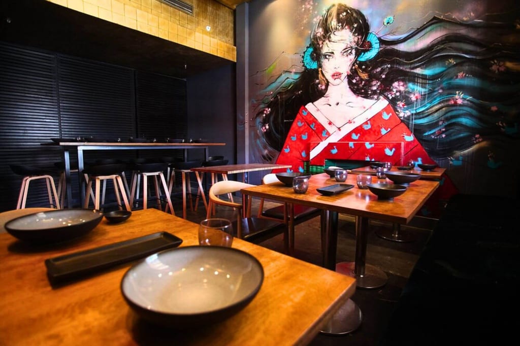 Innenraum eines Restaurants mit gedeckten Holztischen und großem Wandgemälde einer Frau im Kimono.