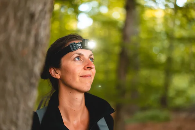 Frau mit eingeschalteter Stirnlampe im Wald, grünes Blattwerk im Hintergrund.