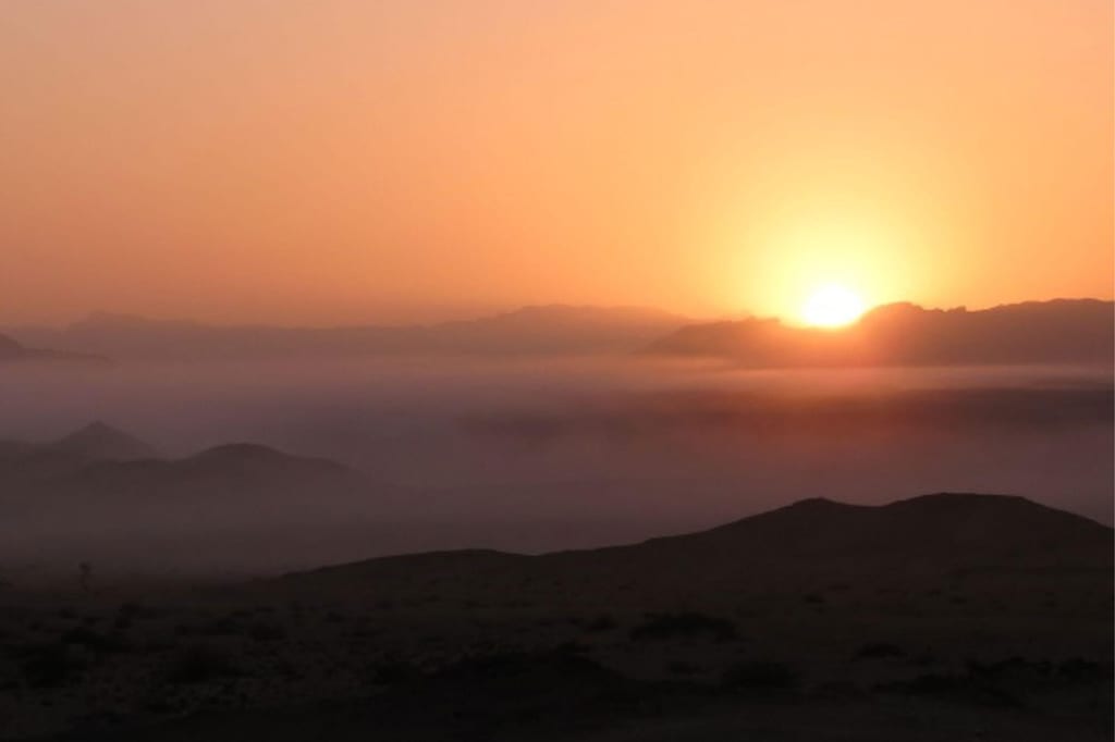 Wüstenlandschaft mit Dämmerungsnebel und tief stehendem orangefarbenen Sonnenball am Horizont
