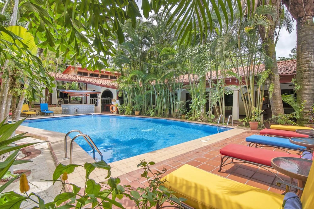 Rechterckiger Pool in einem tropischen Innenhof mit bunten Liegen und Palmen, Tageslicht am Mittag.