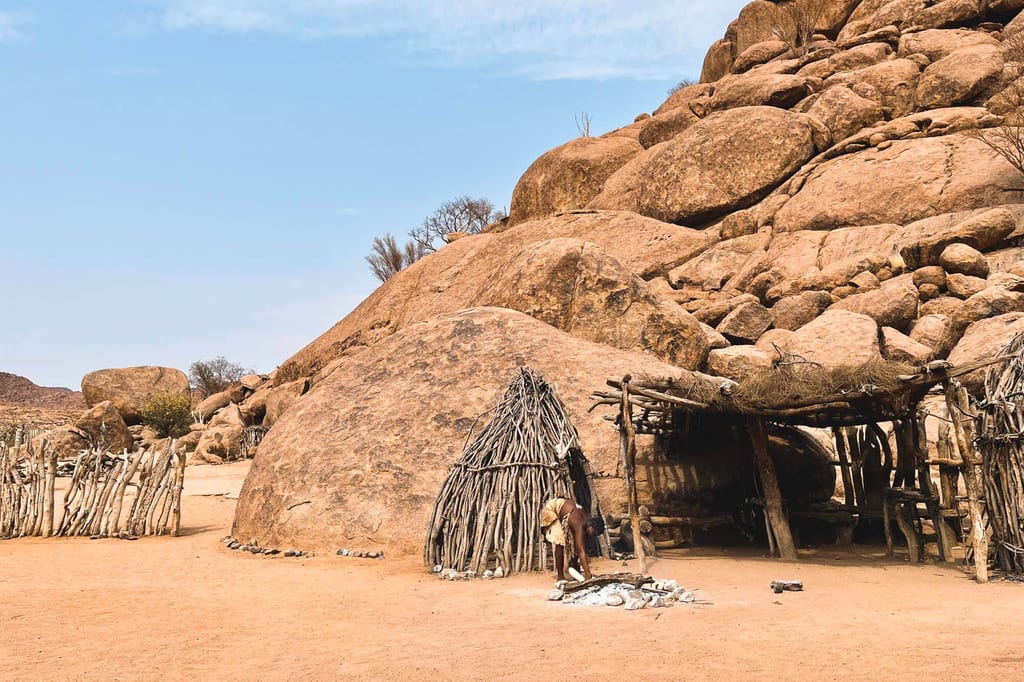 Traditionelle Holzhütten vor großen Felsformationen in trockener, sandiger Landschaft mit einer arbeitenden Person.
